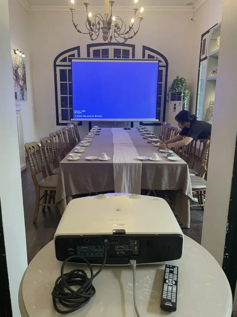 Phòng họp trang bị máy chiếu Epson và bàn dài với ghế gỗ, đang chuẩn bị cho buổi họp hoặc sự kiện