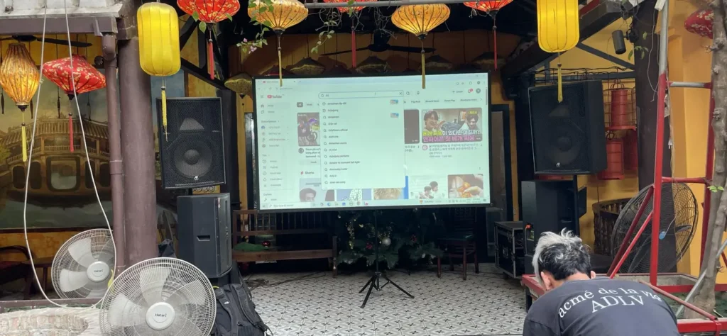 Không gian chiếu phim YouTube với đèn lồng và âm thanh chất lượng Người ngồi trước màn hình chiếu YouTube trong không gian có đèn lồng truyền thống và quạt điện