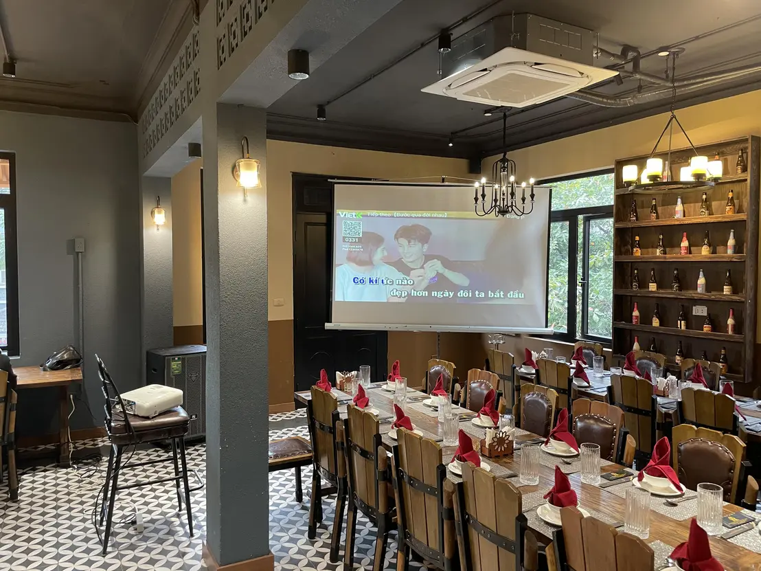 A medium-sized projection screen displaying Vietnamese lyrics in a restaurant or event space with wooden chairs, tables, and decorative lighting. The screen appears to be mounted on a wall, and the setup is suitable for presentations or entertainment.